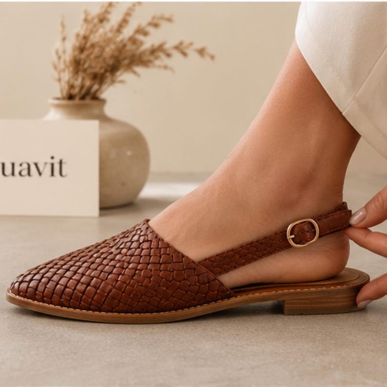 May include: Brown woven leather flat sandal with a slingback strap and a gold-coloured buckle. The sandal has a low heel and a rounded toe. The word "uavit" is visible on a white card in the background.
