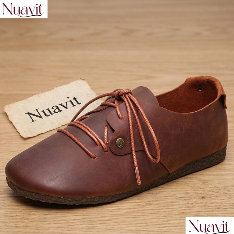 May include: Brown leather lace-up shoe with a rounded toe and a textured sole. The shoe features a decorative button and laces. The brand name "Nuavit" is visible on a tag and in the corner. The shoe is on a wooden surface.