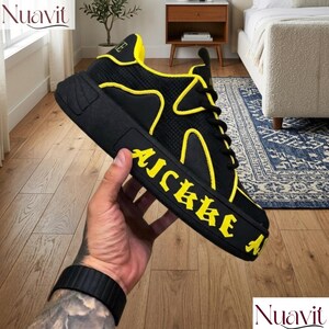 May include: Black and yellow sneaker with the word "AICKKE" in yellow lettering on the side. The shoe has a thick sole and yellow accents. The shoe is held by a person.