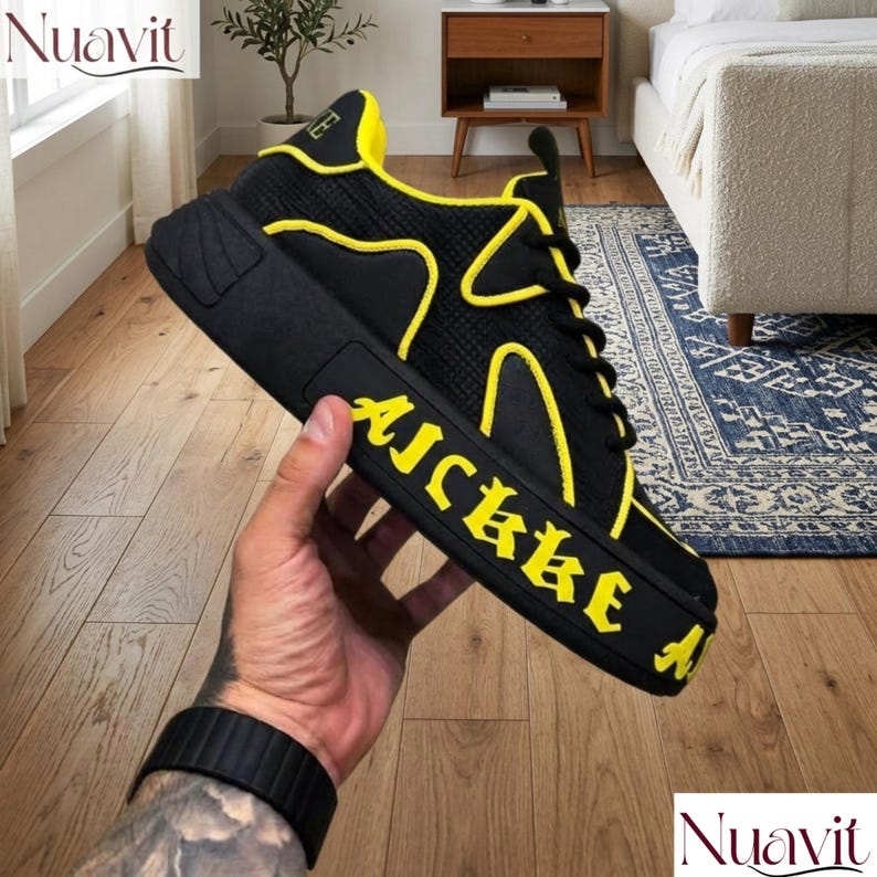 May include: Black and yellow sneaker with the word "AICKKE" in yellow lettering on the side. The shoe has a thick sole and yellow accents. The shoe is held by a person.