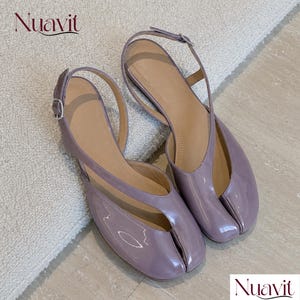 May include: A pair of glossy, mauve-colored split-toe flats with a slingback strap and a silver-toned metal buckle. The shoes feature a distinctive toe separation design and a smooth, shiny finish. The brand name Nuavit is visible in the corner.