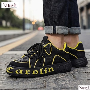 May include: Black sneakers with yellow accents and the word "Carolin" in yellow on the sole. The shoes have a patterned design on the upper and black laces. The brand name "Nuavit" is visible in the top left and bottom right corners.