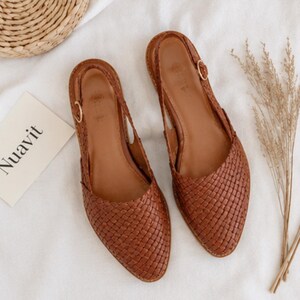 May include: A pair of brown woven leather slingback flats. The shoes have a pointed toe and a small gold buckle on the ankle strap. The word "Nuavit" is printed on a white card next to the shoes.