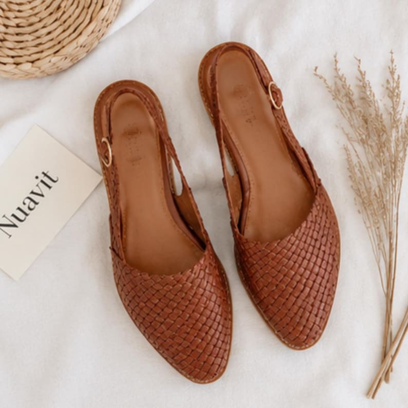 May include: A pair of brown woven leather slingback flats. The shoes have a pointed toe and a small gold buckle on the ankle strap. The word "Nuavit" is printed on a white card next to the shoes.