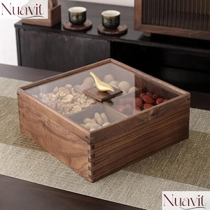 Puede incluir: Una caja de madera para servir con tapa de cristal, dividida en cuatro compartimentos. La caja está llena de frutos secos y fruta deshidratada. Un asa dorada con forma de pájaro se encuentra sobre la tapa de cristal. La caja está sobre una mesa con un camino de mesa estampado.
