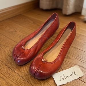 May include: A pair of red leather ballet flats with a split toe design. The shoes have a rounded toe and a small decorative detail at the vamp. The brand name "Nuavit" is on a small tag.
