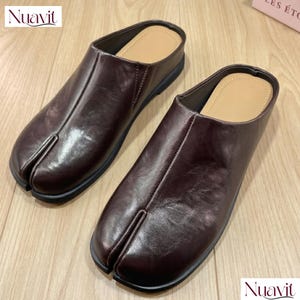 May include: A pair of dark brown slip-on mules featuring a distinct split-toe design. The shoes have a smooth, polished finish, a central seam running down the vamp, and a tan-coloured interior footbed. The soles are black and flat.