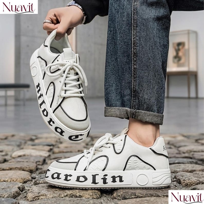 May include: White sneakers with black accents and the word "carolin" printed on the side. The shoes have white laces and a thick sole. The word "nildora" is printed on the side of the other shoe. The shoes are being held up by a person.
