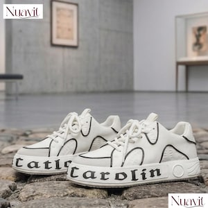 May include: Pair of white sneakers with black trim and the word "Carol&iacute;n" printed on the side. The shoes have white laces and a textured design on the upper. The brand name "Nuavit" is visible in the corner.