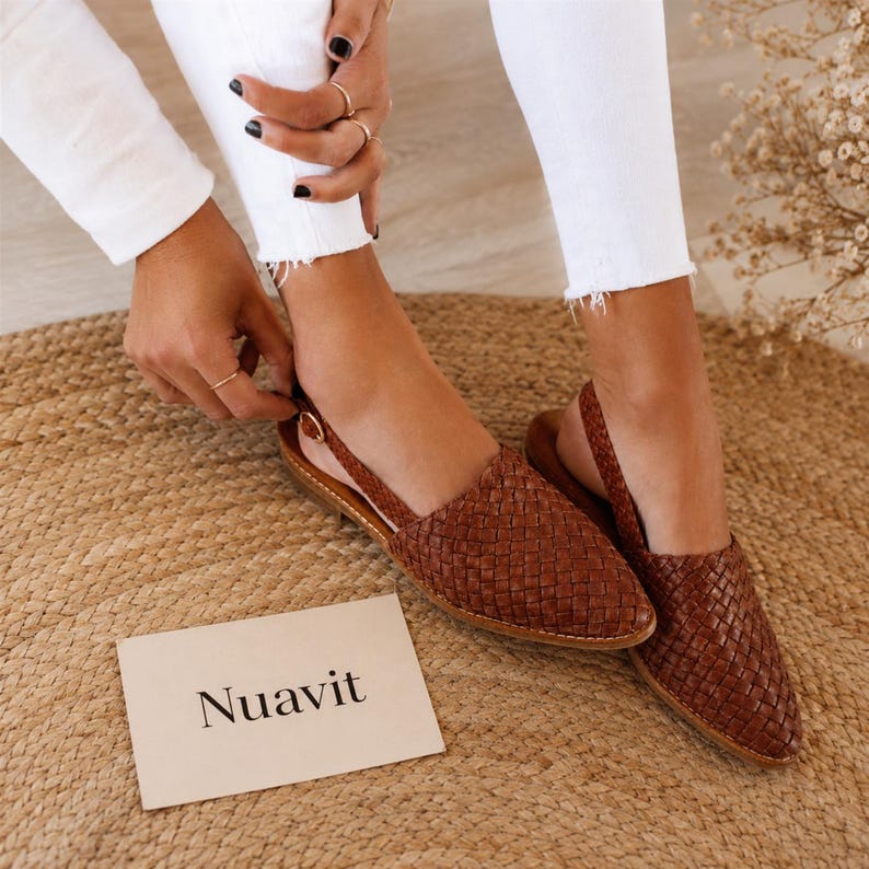 May include: Brown woven leather slingback flats with a pointed toe. The shoes have a strap around the ankle and are displayed on a woven mat. The word "Nuavit" is printed on a white card.
