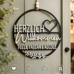 Könnte beinhalten: Ein rundes schwarzes Wandschild mit dem weißen Text "HERZLICH Willkommen" und "FELIX, FABIAN & NICOLE", einem Herzumriss und einem Blattzweig-Design. Das Schild ist an einem Juteseil mit zwei Holzperlen an der Oberseite aufgehängt.