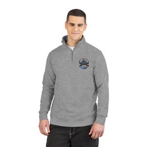 May include: A gray quarter-zip sweatshirt with a small embroidered emblem on the left chest. The emblem features a stylized cityscape with blue and red accents above blue waves. The sweatshirt has a mock neck and a silver zipper.