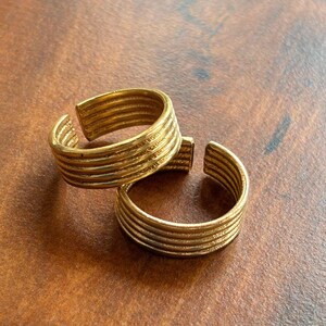 May include: Two gold-colored, adjustable rings. Each ring features a series of horizontal ridges, creating a textured surface. The rings are open at the front, allowing for size adjustment. The rings are displayed on a brown surface.