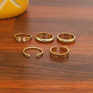 May include: A set of five gold-toned rings. One ring features two small clear stones at the opening, one has a dotted texture, one is a plain band, one has a simple open design with small spheres, and one displays engraved celestial symbols.