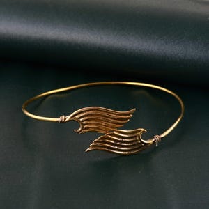 May include: A gold-toned metal bangle bracelet featuring two symmetrical wing-shaped charms that meet in the center. The thin, circular band is connected to each wing with small wire-wrapped details, creating a cohesive and delicate jewelry piece.