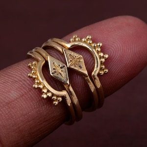 Può includere: Un set di due anelli color oro caratterizzati da un design ad arco curvo con piccole perline sferiche lungo il bordo esterno. Ogni anello presenta una placca centrale a forma di diamante incisa con un motivo a stella, creando un aspetto a strati.