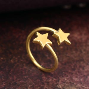 Può includere: Un anello color oro con due decorazioni a forma di stella. Le stelle sono posizionate su entrambi i lati dell'apertura dell'anello, creando un design celeste unico. L'anello è realizzato in materiale metallico lucido.