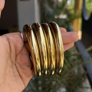 May include: A set of four thick, polished gold-tone metal bangle bracelets with a smooth, rounded surface. The bangles are stacked together, showcasing a reflective, high-shine finish that catches the light across their curved, cylindrical forms.