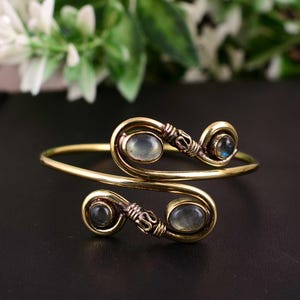 May include: A gold-toned cuff bracelet with three oval, iridescent gemstones. The bracelet features a swirling design with decorative wire detailing around the gemstones. The bracelet is displayed on a dark surface.