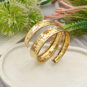 May include: A gold-toned metal cuff bracelet featuring a spiral design with a hammered, textured surface. The bracelet has an open-ended construction that wraps around itself, creating a layered appearance with a polished, reflective finish.