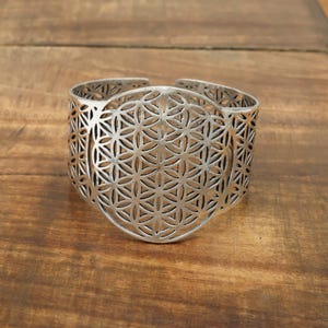 Könnte beinhalten: Silberfarbenes Armreif-Armband mit offenem Design. Das Armband hat ein zentrales kreisförmiges Muster aus miteinander verbundenen blütenartigen Formen, umgeben von einem geometrischen Gitter. Das Armband wird auf einer Holzoberfläche präsentiert.