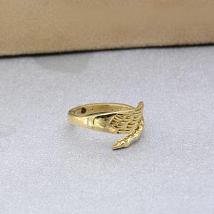 Op de afbeelding: Een goudkleurige ring met een vleugelontwerp. De ring wikkelt zich om de vinger, met het vleugeldetaill op één kant. De ring is gemaakt van een glanzend, metalen materiaal.