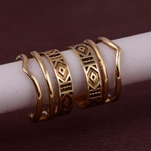 Könnte beinhalten: Ein Set aus fünf goldfarbenen Ringen, die auf einem weißen zylindrischen Ständer angeordnet sind. Zwei Ringe haben ein glattes, gewelltes Banddesign, während die anderen drei Ringe ein geometrisches Muster aus Rauten und Punkten aufweisen.