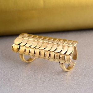 Pode incluir: Um anel dourado com um design único. O anel apresenta uma série de elementos sobrepostos, em forma de escamas, que formam uma forma retangular longa sobre o dedo. O anel tem duas bandas para os dedos.