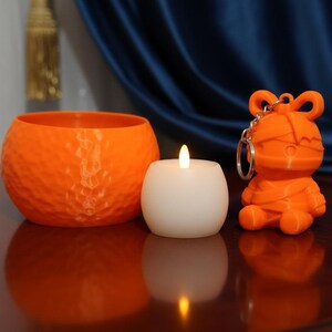 May include: An orange, textured candle holder and a white candle with a flickering flame sit next to an orange keychain shaped like a cartoon character. The items are on a reflective surface.