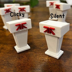 May include: White podiums with red accents and the words "Clicky" and "Silent" printed on them. The podiums have a rectangular top and a trapezoidal base. The background is a dark wooden surface.