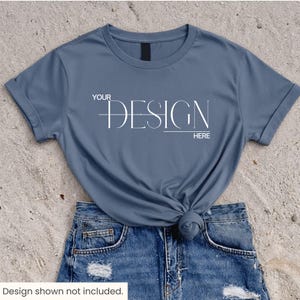 Peut inclure: Un t-shirt bleu ardoise à manches courtes avec un col rond et des manches retroussées. Le centre du t-shirt présente un texte blanc indiquant YOUR DESIGN HERE avec une ligne horizontale traversant le mot DESIGN. Le t-shirt est rentré dans un short en jean bleu.
