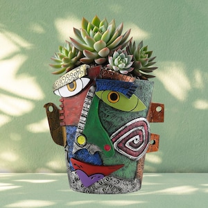 May include: A decorative planter shaped like a stylized human face with a multi-colored, textured surface featuring red, green, blue, and silver sections. A cluster of green succulent plants grows from the top of the head-shaped container.
