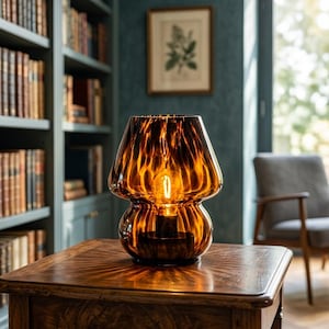 May include: A mushroom-shaped table lamp made of translucent amber-colored glass with a dark, swirling tortoiseshell pattern. The lamp features a wide, rounded shade and a bulbous base, emitting a warm, glowing light from an internal filament bulb.