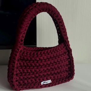 Puede incluir: Un bolso de mano de ganchillo burdeos con un asa redondeada. El bolso tiene forma rectangular y un aspecto texturizado. Una pequeña etiqueta blanca con la palabra "Mapsi" está adherida a la parte delantera del bolso. El bolso está sobre una superficie blanca.