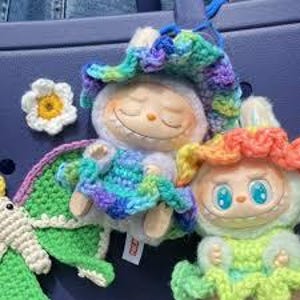 May include: A selection of vibrant crochet dolls and accessories. The dolls have whimsical designs with closed eyes and smiling faces. One doll is dressed in a blue, green, and purple hat and outfit. A white and yellow flower and a green and white creature are also present.