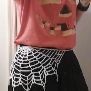 May include: A coral-colored t-shirt featuring a dark brown jack-o'-lantern face graphic, paired with a white crocheted spiderweb-patterned waist accessory that hangs over a solid black skirt, finished with small dangling clear beads at the bottom.
