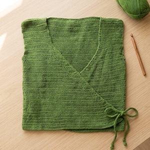 May include: A handmade, green crochet wrap top with a V-neck and tie closure. A ball of green yarn and a crochet hook are also visible. The top is made of a soft, textured yarn, perfect for a summer wardrobe.