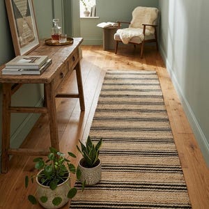 Könnte beinhalten: Ein Flur mit einem Naturfaserteppich mit schwarzen Streifen. Der Teppich liegt auf einem Holzboden. Auf der linken Seite befindet sich ein Holztisch mit Büchern und einem gerahmten Kunstwerk. Im Hintergrund ein Stuhl mit Schaffell und ein runder Spiegel.