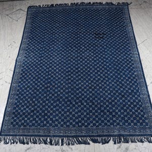 Puede incluir: Una alfombra rectangular con una base de color azul índigo profundo y un patrón floral geométrico blanco repetitivo en el centro. La alfombra está enmarcada por un borde decorativo y terminada con flecos cortos y anudados en los extremos.