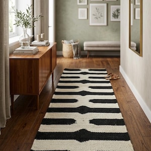 May include: A black and white striped runner rug in a hallway. The rug has a natural fiber border and is placed on a hardwood floor. The hallway also features a wooden cabinet, a mirror, and framed botanical prints.