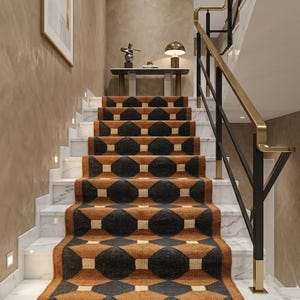 Könnte beinhalten: Eine Treppe mit einem gemusterten Teppichläufer. Der Läufer weist ein sich wiederholendes geometrisches Design in Schwarz, Braun und Creme auf. Die Treppe ist aus Marmor und die Wände sind in einem warmen Neutralton gehalten. Ein goldfarbener Handlauf verläuft entlang der Seite.