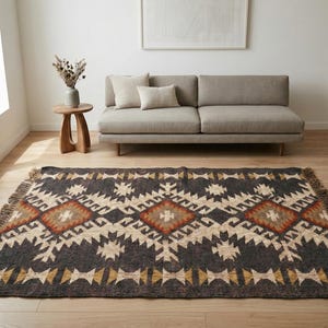 Op de afbeelding: Een rechthoekig vloerkleed met een geometrisch patroon in grijstinten, beige, oranje en geel. Het vloerkleed heeft een gefranjerde rand en ligt op een houten vloer. Een bank en een bijzettafel staan op de achtergrond.