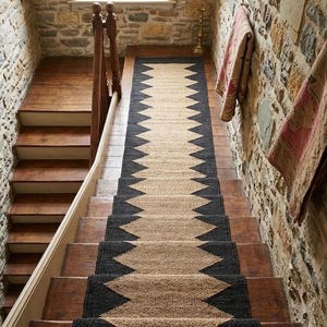 Puede incluir: Una alfombra para escaleras con un diseño geométrico en negro y beige. La alfombra se coloca sobre escaleras de madera, con paredes de piedra a ambos lados. El patrón en zigzag de la alfombra contrasta con los escalones de madera. Las escaleras conducen a una ventana.