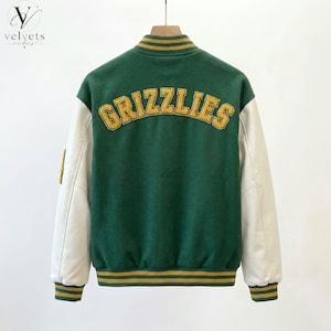 May include: Green and cream varsity jacket with the word "GRIZZLIES" in gold lettering across the back. The jacket has cream-colored sleeves and gold and green striped trim on the collar, cuffs, and hem. The jacket is hanging on a wooden hanger.