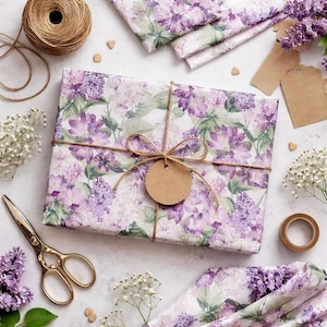 May include: A rectangular gift box wrapped in paper featuring a purple and green lilac floral pattern. The box is tied with brown twine in a bow, holding a blank circular brown paper gift tag. Nearby are gold scissors, twine, and lilac flowers.