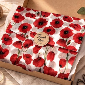 May include: A cardboard shipping box containing items wrapped in white tissue paper featuring a vibrant red poppy flower pattern. The paper is secured with a round, light brown sticker printed with the text Thank You and a small heart symbol.