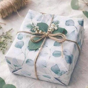 May include: A square gift box wrapped in white paper featuring a pattern of green eucalyptus leaves. The package is tied with brown twine and decorated with a small sprig of fresh green eucalyptus leaves and tiny white flowers on top.