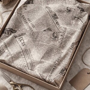 May include: A rectangular cardboard box filled with off-white tissue paper printed with black vintage newspaper text, including headlines like Art and Entertainment and Irish Press, with a spool of twine and metal scissors nearby.