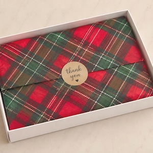 May include: A rectangular white cardboard box containing an item wrapped in red, green, and brown plaid tissue paper. A circular tan sticker is placed in the centre of the paper, featuring the black text thank you and a small heart symbol.