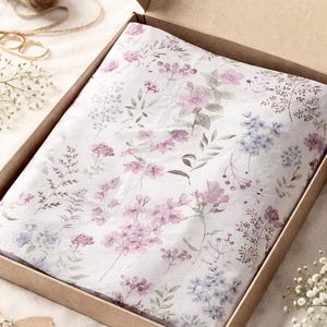 May include: A rectangular cardboard shipping box containing white tissue paper printed with a delicate watercolour pattern of pink and blue wildflowers, small green leaves, and thin stems, neatly folded to fit inside the open container.